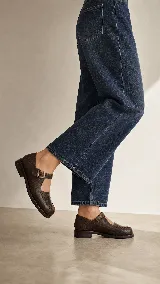 Mocasín de cuero color marrón con acabado texturizado, diseño de punta redonda con costuras artesanales visibles y cierre mediante una tira con hebilla lateral.