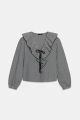 Blusa de manga larga con estampado de cuadros vichy en blanco y negro. Presenta un escote en V adornado con un volante amplio y un lazo decorativo en el centro. Las mangas terminan en puños elásticos.