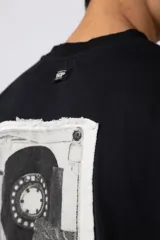 Remera negra de corte oversize confeccionada en tela pesada. Presenta un bordado frontal con la palabra 'REWIND' y un diseño de parche en la espalda con estética de papel roto.