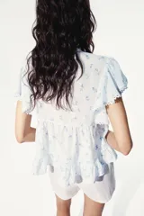 Blusa celeste claro con delicado estampado floretado azul, escote en V pronunciado, mangas cortas tipo mariposa y cierre frontal con botones. Presenta inserciones de encaje y alforzas decorativas en la parte delantera, además de un dobladillo con volados y detalles de encaje.