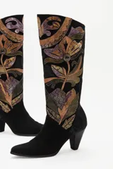 Bota de caña alta confeccionada en gamuza negra, con un diseño artesanal de parches en cueros texturizados y metalizados con motivos florales. Presenta un taco cónico y punta almendrada.