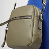 Bandolera de diseño compacto en color verde oliva, con cierre superior y bolsillo frontal con cierre. Incluye una correa ajustable de tela en color crudo con detalles en verde.