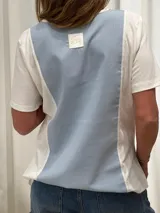 Remera de diseño combinado en algodón y lino, con panel frontal y posterior en color amarillo suave y laterales en blanco. Presenta detalles de puntilla vertical en la unión de los paneles, cuello redondo y un corte asimétrico con el ruedo trasero ligeramente más largo.