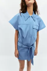 Vestido corto de estilo camisero color azul, con cuello solapa, manga corta y cierre frontal con botones. Presenta un bolsillo de plastrón con solapa en el pecho y un cinturón ajustable del mismo tejido en la cintura.