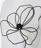 Remera de corte box, levemente amplia, confeccionada en algodón. Presenta cuello redondo, mangas cortas y un diseño de flor bordada en hilo negro en la parte delantera.
