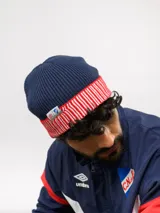 Gorro tipo beanie de punto acanalado en color azul marino, con vuelta en color rojo y blanco a rayas. Incluye etiqueta lateral con el logo de Umbro y el escudo del Club Nacional de Football.