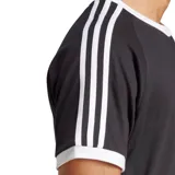 Remera negra de corte clásico con cuello redondo y mangas cortas con ribete blanco. Presenta las tres rayas blancas características de Adidas en los hombros y el logo del trifolio bordado en blanco en el pecho.