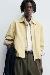 Cazadora de tejido con algodón, color amarillo pálido, con cuello solapa y cierre frontal de cremallera. Presenta bolsillos frontales de vivo y bajo ajustable con elástico en los laterales.