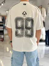 Remera regular color off-white, con diseño gráfico en la espalda que incluye el número 99 en gran tamaño y detalles en negro. Cuenta con ribetes negros en las costuras de los hombros y mangas.