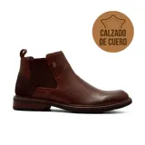 Bota tipo Chelsea de cuero color marrón, con paneles laterales elásticos, detalle de remache metálico y refuerzo de gamuza en el talón. Cuenta con suela de goma con textura y tirador trasero.