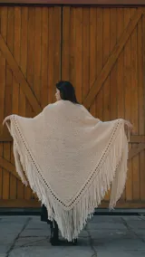 Ruana artesanal tejida en lana merino color crudo, con diseño de punto abierto en los bordes y flecos largos en todo el contorno.