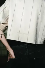 Camiseta crop de manga corta con cuello polo, confeccionada en tela técnica tipo dry fit. Presenta un diseño a rayas verticales finas en color beige, con paneles laterales de malla en tono camel y detalles de parches bordados en el frente.