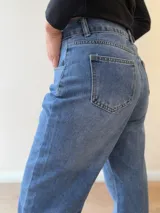 Pantalón de jean corte mom fit con tiro alto, confeccionado en denim de lavado clásico. Presenta cierre frontal con botón y cierre metálico, con bolsillos funcionales delanteros y traseros.