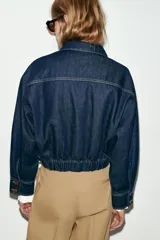 Chaqueta estilo bomber confeccionada en tejido denim azul oscuro. Presenta cuello solapa, cierre frontal con cremallera, mangas largas y bajo elástico. Incluye bolsillos delanteros de plastrón con solapa y botones.