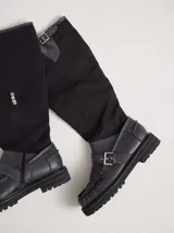 Bota híbrida de caña alta confeccionada en lona negra en la parte superior y cuero negro en la base tipo mocasín, con plataforma, suela track y tira con hebilla metálica cruzada en el empeine.