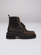 Borcego de gamuza color chocolate con paneles laterales elásticos negros y suela track dentada. Presenta sistema de cordones con ganchos metálicos y tirador en la parte trasera.