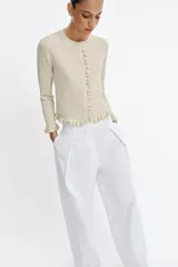 Chaqueta de punto canalé en color beige con hilo metalizado, cuello redondo, manga larga y cierre frontal con botones. Presenta detalles de volantes en la tapeta de los botones, el ruedo y los puños.