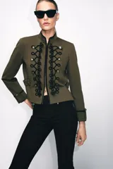 Blazer de estilo militar confeccionado en algodón, con cuello subido y manga larga. Presenta un diseño estructurado con detalle de alamares negros a contraste en el frente, botones metálicos decorativos y trabillas laterales. Cuenta con cierre frontal oculto y forro interior.