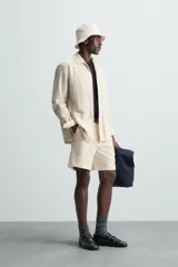 Sobrecamisa de hombre de corte relajado, color beige claro con finas rayas verticales en tono más oscuro. Presenta cuello bowling, manga larga con puño abotonado, bolsillos de parche en el pecho y en la cadera, y aberturas laterales en el bajo.