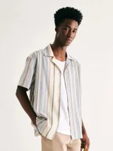 Camisa de manga corta con cuello cubano, confeccionada en tejido de lino a rayas verticales en tonos beige, azul y naranja. Presenta un corte holgado y cierre frontal con botones.