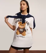 Remera blanca de algodón con cuello redondo y manga corta. Presenta un estampado central de un oso de peluche marrón vistiendo un buzo negro con el texto "NEW YORK" en blanco.