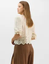 Blusa de algodón semitransparente con diseño floral bordado a tono. Presenta escote en V, cierre frontal con botones y mangas al codo ligeramente abullonadas. Incluye detalles de encaje calado estilo macramé en los puños y en el ruedo con terminaciones en ondas.