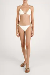 Calzón de bikini color crema con tiras laterales ajustables y detalles de cuentas metálicas doradas en los extremos.
