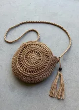 Bolso redondo tejido a mano en fibra natural (papel y algodón), color marrón claro, con forro interior. Posee correa trenzada larga y un adorno de borlas con cuenta de madera oscura.