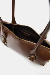 Bolso de hombro con acabado efecto craquelado en color marrón oscuro. Presenta doble asa de hombro, cierre superior con cremallera y detalles metálicos dorados en la parte frontal y laterales.