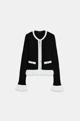 Chaqueta de punto acanalado en color negro con detalles en blanco en cuello, tapeta de botones, bolsillos y volados en el ruedo y puños. Presenta cierre frontal con botones dorados.