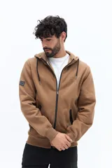 Campera de felpa color camel con cierre frontal, capucha ajustable con cordones negros y bolsillos laterales con cierre negro. Presenta un estampado pequeño en el pecho con texto en color oscuro.