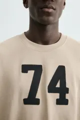 Sudadera de manga corta color beige, corte relajado (relaxed fit) con cuello redondo y número "74" bordado en negro en el frente.