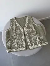 Chaleco marrón con bordados florales en color crema en los bordes y el frente. Presenta un diseño sin mangas con escote en V.