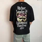 Remera negra de corte oversize con estampa gráfica en la espalda que presenta un oso con anteojos y bandana, acompañada de la frase 'Richer, Louder & Unbothered Than Ever'.
