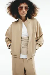 Cazadora bomber de color beige con cuello subido, manga larga y bolsillos delanteros. Cuenta con cierre frontal de cremallera metálica y cordón ajustable en el dobladillo.