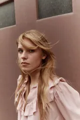 Blusa rosada de manga larga con cuello alto y lazo para atar. Presenta ribetes de volados que cruzan el pecho y puños con elástico fruncido.