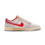 Championes Nike Dunk Low con diseño de paneles en tonos gris claro y blanco, destacando el swoosh lateral y detalles en el talón y cuello en color rojo vibrante. Presenta cordones en tono crema y una suela con efecto envejecido.