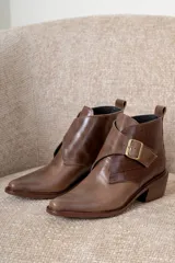 Botin de cuero color bordeaux con diseño de puntera fina y taco cuadrado. Presenta una solapa lateral con hebilla metálica dorada y detalle de textura tipo reptil en la parte posterior.