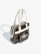Bolso tipo shopper de Adidas, confeccionado en material sintético con acabado de charol brillante en color marrón oscuro y detalles en blanco. Presenta el logo del Trifolio estampado en el frente, bolsillo exterior con cierre y asas largas para llevar al hombro.