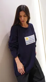 Sweater de hilo de algodón color verde militar, con cuello redondo y manga larga. Presenta un diseño tejido de la bandera uruguaya en el frente.