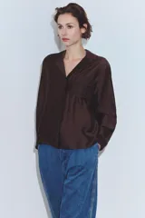 Camisa de manga larga color marrón oscuro, con cuello solapa, escote pico y bajo asimétrico. Presenta un bolsillo plastrón en el delantero con bordado combinado a tono.