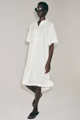 Vestido camisero de lino con cuello solapa, manga corta y cierre frontal con botones. Presenta un diseño con bajo redondeado y aberturas laterales.