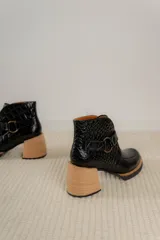 Bota corta de cuero vacuno con textura de reptil, color marrón oscuro. Cuenta con un taco cuadrado negro, suela de plataforma y una tira decorativa con hebilla metálica circular en el tobillo.