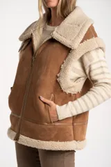 Chaleco estilo gamulán de corte oversize, confeccionado en material tipo gamuza color camel con interior y bordes de corderito sintético color crema. Presenta cierre frontal completo, bolsillos laterales y detalles de tiras decorativas en los hombros.