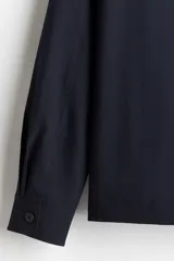 Sobrecamisa de tejido suave color azul marino, con cuello inglés, tapeta lisa y botones frontales. Presenta dos bolsillos superiores abiertos y canesú en la espalda.