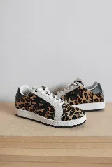 Zapatillas urbanas con capellada de cuero blanco, suela de caucho y cordones blancos. Presentan detalles en gamuza gris claro en los laterales y puntera, y un parche negro con un logo de corona en animal print en el lateral. El talón es de cuero verde.