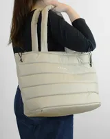 Bolso tipo tote color beige con diseño acolchado y asas tubulares. Presenta el logo de la marca en el centro.