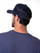 Gorro trucker azul marino de siete paneles, con visera curvada y parte trasera de malla transpirable. Cuenta con un parche bordado en rombo con el logotipo de Caterpillar en el frente y cierre ajustable a presión en la parte posterior.