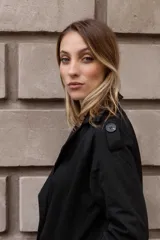 Trench clásico de largo midi en color beige, con diseño de doble abotonadura, solapas amplias, detalles en hombros y correas ajustables en los puños.