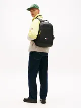 Mochila negra de poliuretano con compartimento principal y cierre de doble cremallera con tiradores amarillos a contraste. Cuenta con bolsillo frontal con cremallera, asa superior, correas ajustables y acolchadas, y parche engomado de Tommy Jeans en la parte delantera.